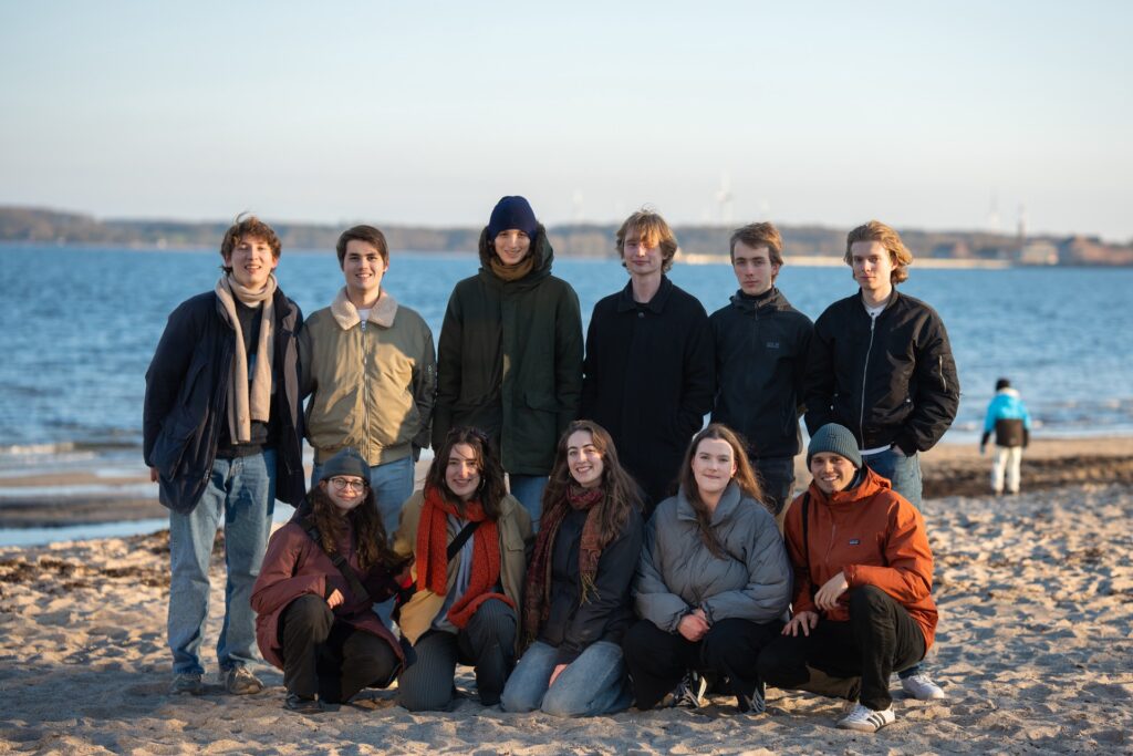 Eine Gruppe von 11 Personen die in zwei Reihen an einem Strand stehen. Im Hintergrund sieht man das Meer.